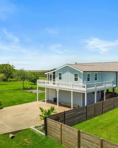 White cottage with covered porch, two blue chairs, large shade trees, and small shed in grassy yard